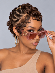 A close-up of a model wearing chunky, rhinestone-embellished cat-eye sunglasses with a gold-tone circular logo on the frame. The model has styled brown curly hair, gold hoop earrings, and is wearing a beige one-shoulder top against a solid gray studio background.