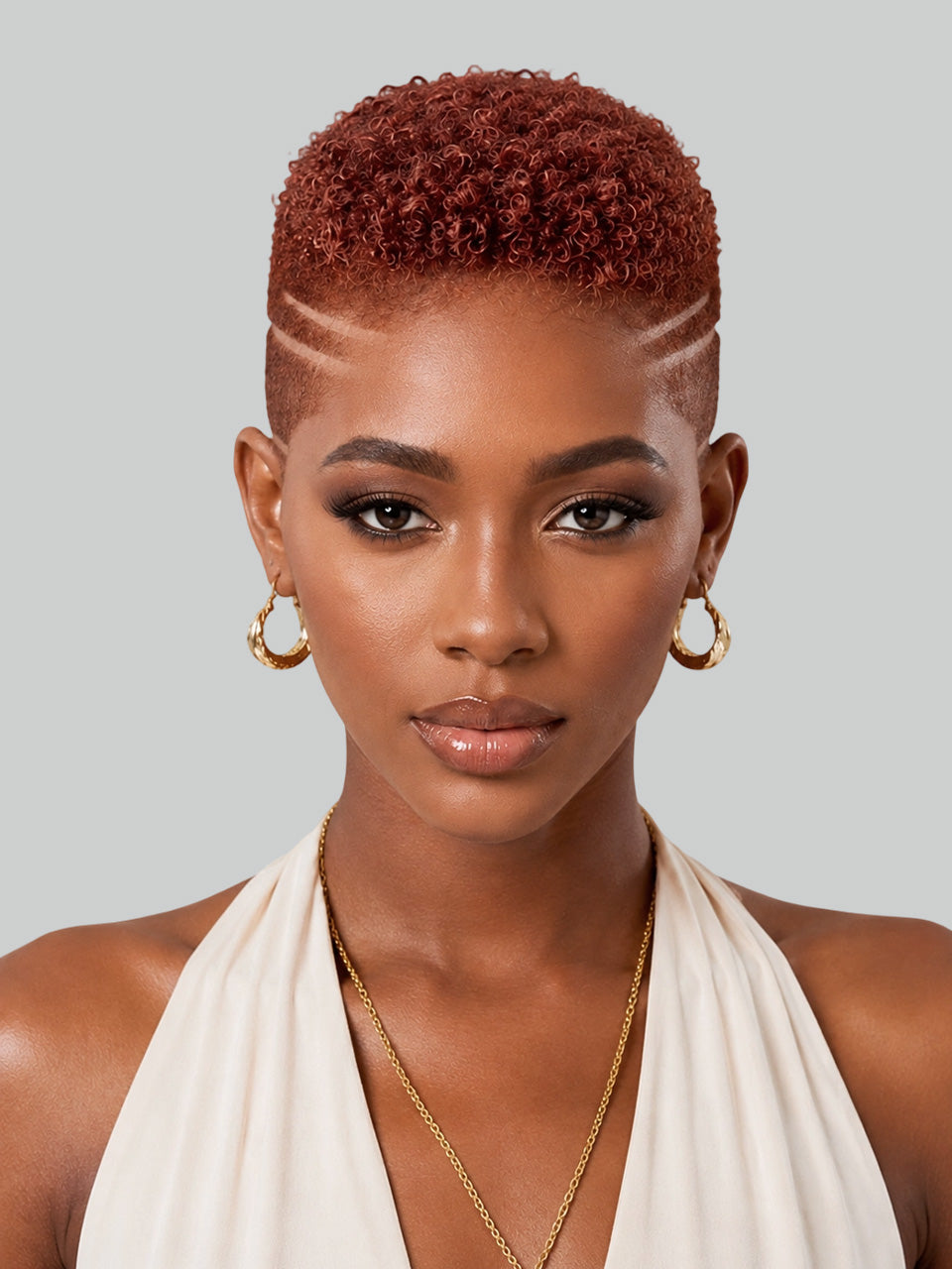 A close-up portrait of a model with a short, textured reddish-brown hairstyle, featuring geometric shaved lines on the sides. She is wearing gold hoop earrings and a gold chain necklace, paired with a cream halter top, set against a solid light gray background.