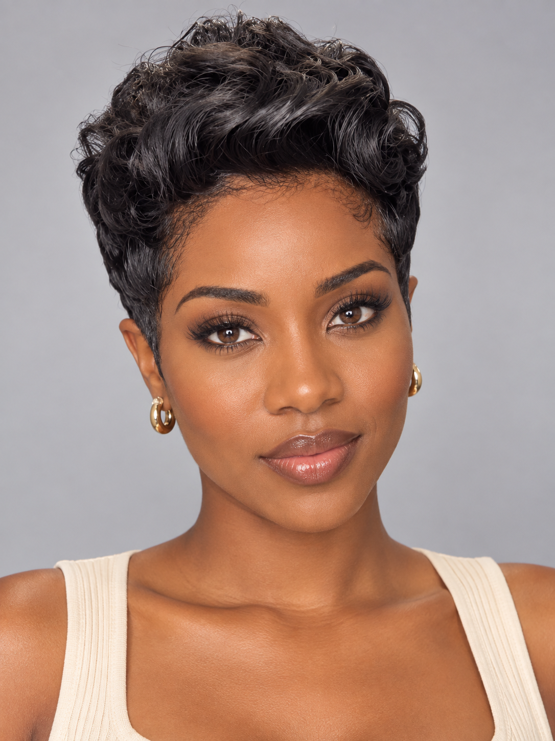 A close-up portrait of a woman with short, curly black hair, wearing gold hoop earrings and a cream ribbed tank top against a solid gray background.