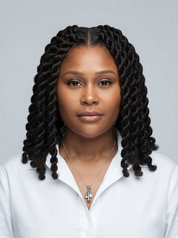 A woman with long, dark box braids or twists wearing a white collared shirt and a silver cross pendant necklace, photographed against a solid light gray background.