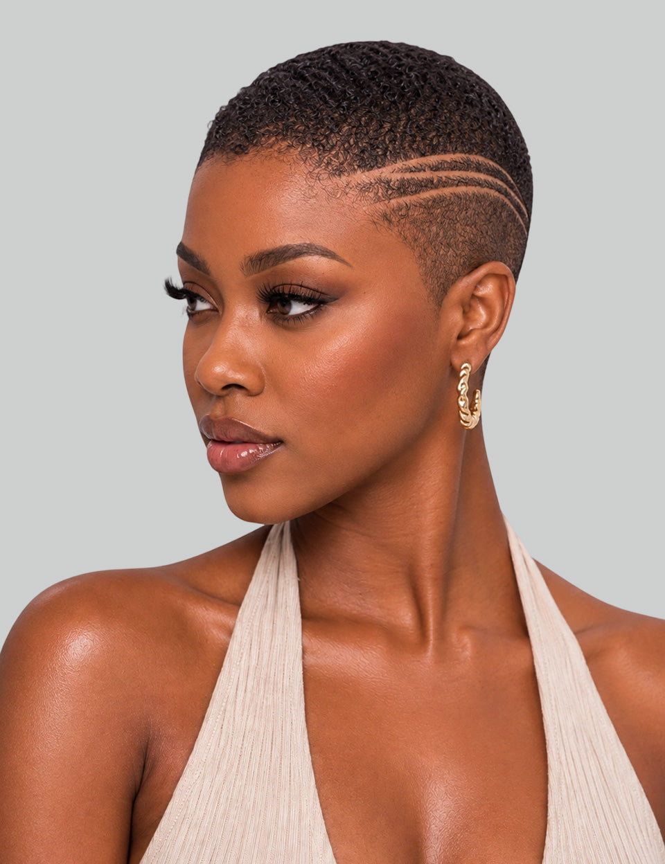 A close-up portrait of a woman with a short, textured black hairstyle featuring three parallel side-combo lines. She is wearing a beige halter top and gold hoop earrings against a solid light gray background.