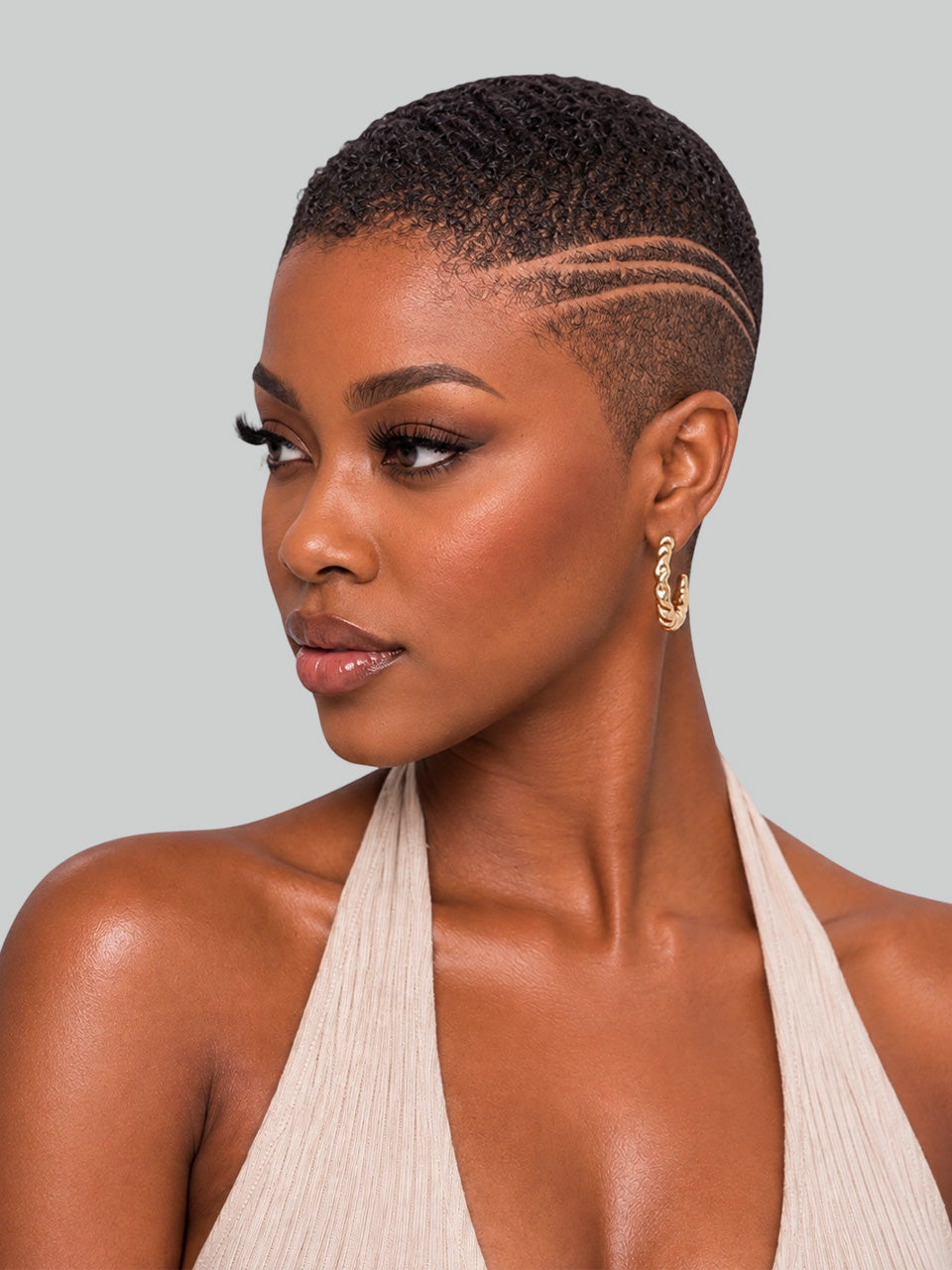 A close-up portrait of a woman with a short, textured black hairstyle featuring three parallel side-combo lines. She is wearing a beige halter top and gold hoop earrings against a solid light gray background.