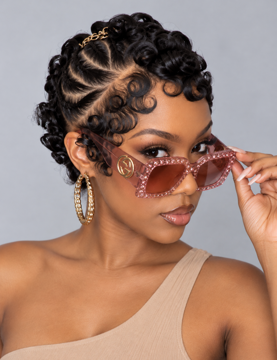 A woman with styled curly hair wears translucent pink rhinestone-trimmed cat-eye sunglasses with gold-tone frames and brown lenses, posed against a plain gray studio background.