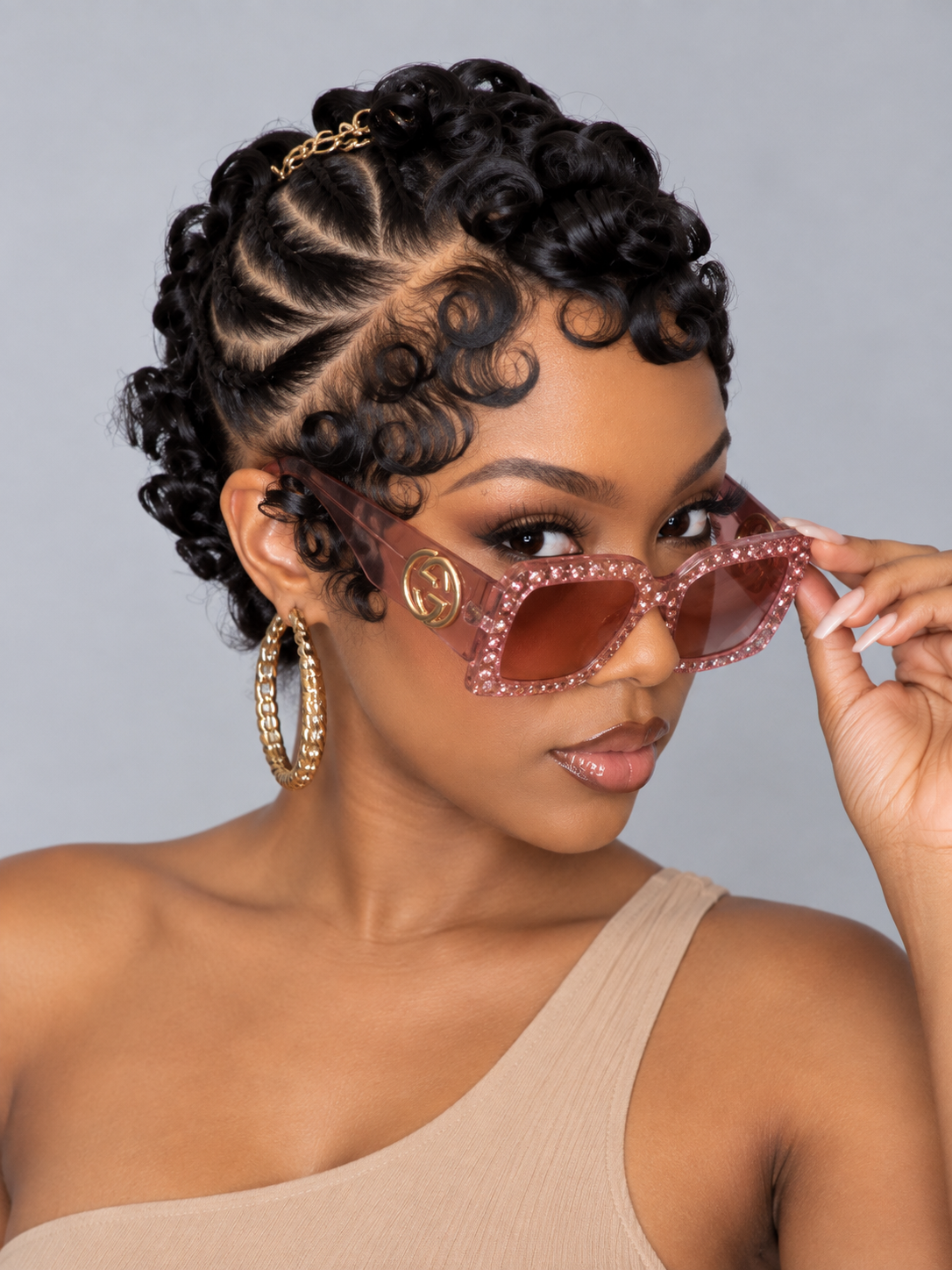 A woman with styled curly hair wears translucent pink rhinestone-trimmed cat-eye sunglasses with gold-tone frames and brown lenses, posed against a plain gray studio background.