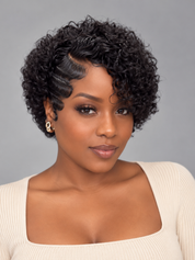 A woman with a short, curly black wig styled with a side part, wearing a cream ribbed top against a solid gray studio background.