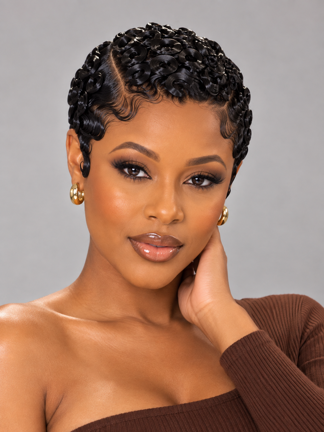 A close-up portrait of a woman with short, tightly curled black hair styled in a braided or twisted updo, wearing gold hoop earrings and a brown ribbed strapless top against a plain gray studio background.