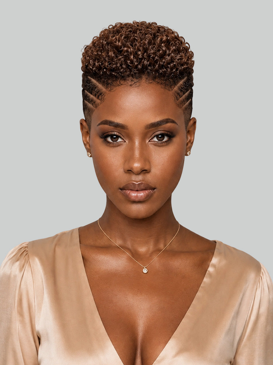 A woman with short, tightly curled brown hair styled in a high puff with side cornrow details, wearing a beige satin top and a delicate gold necklace against a solid gray background.
