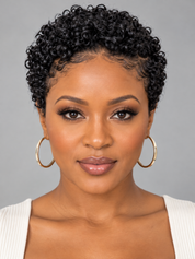 A close-up portrait of a woman with short, tightly curled black hair, wearing large gold hoop earrings and a white ribbed top against a plain gray background.