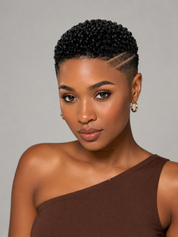 A close-up portrait of a woman with a short, textured black wig and a sharp side undercut, wearing a brown one-shoulder top against a neutral gray background.