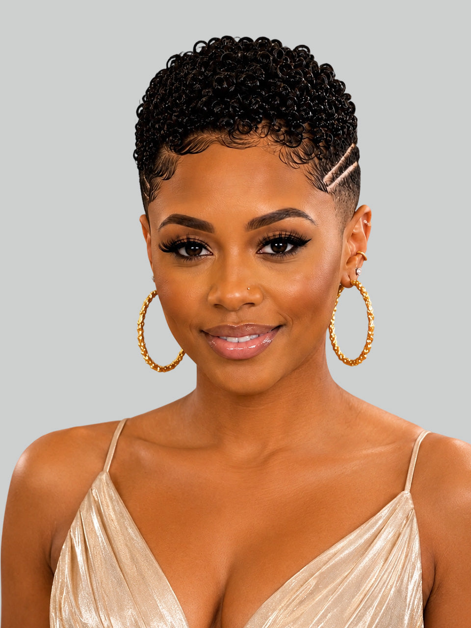 A woman with short, curly black hair styled with a side parting and a small hair clip, wearing large gold hoop earrings and a champagne-colored spaghetti strap top against a solid gray background.