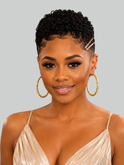 A woman with short, curly black hair styled with a side parting and a small hair clip, wearing large gold hoop earrings and a champagne-colored spaghetti strap top against a solid gray background.