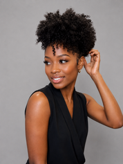 A woman with a voluminous, curly black afro hairstyle wearing a black sleeveless top, posing against a solid gray background.