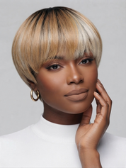A woman with a short, straight, bob-cut wig with bangs in a blonde and dark brown/black ombre style, wearing a white top against a light gray background.