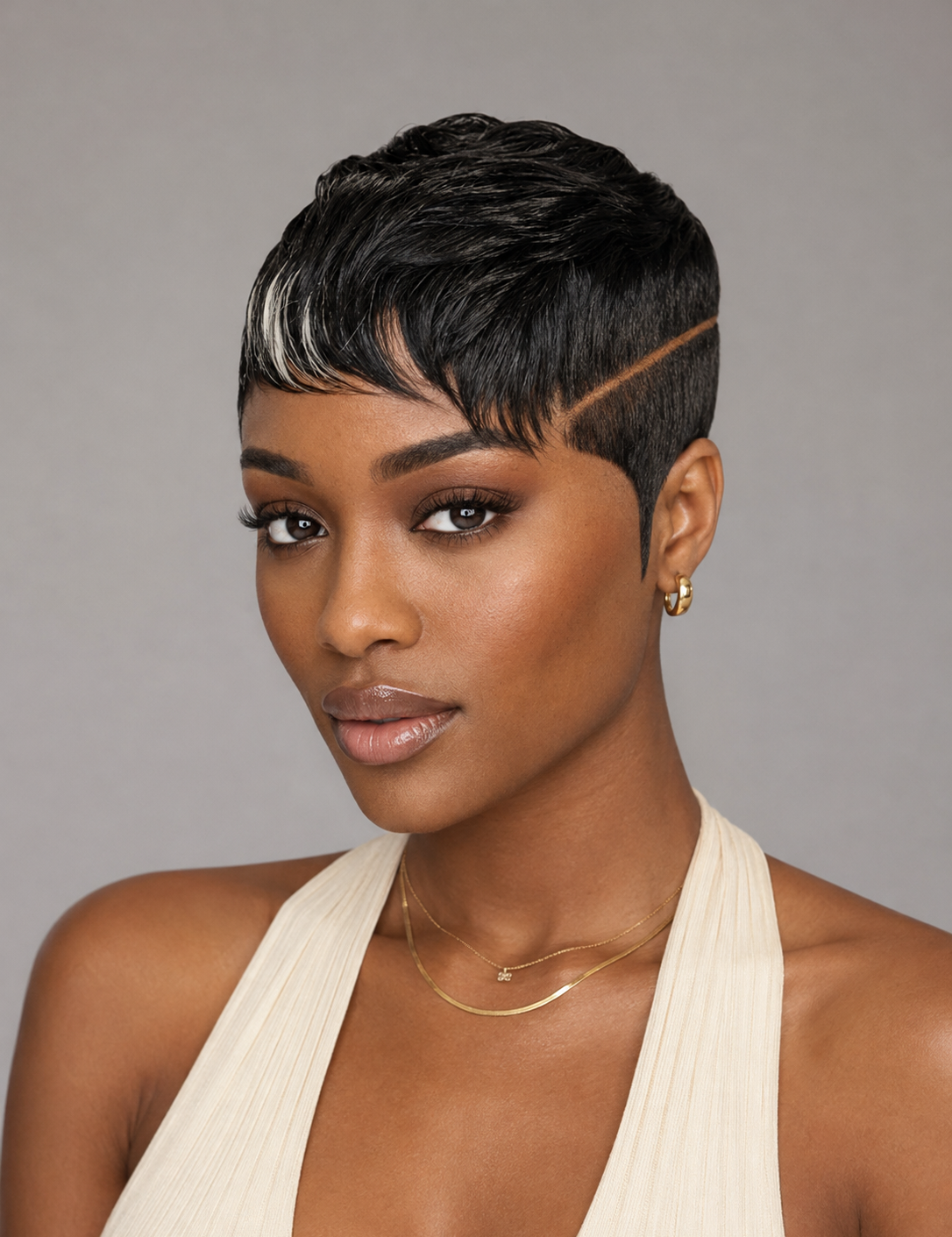 A woman with a short, textured black pixie cut and a side part, wearing a cream halter top and gold jewelry, photographed against a solid gray background.