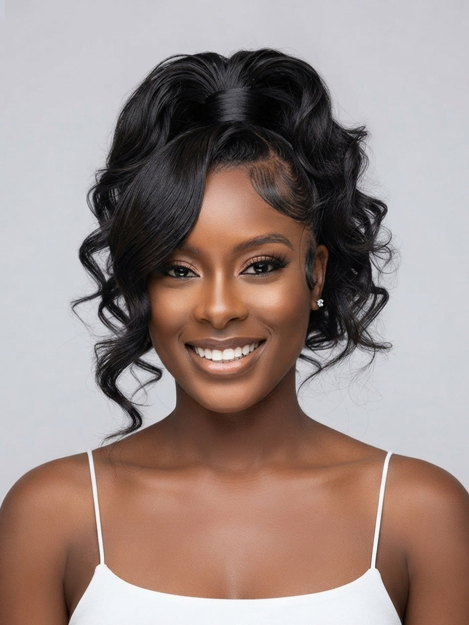 A smiling woman with a high-volume black faux hair extension or wig styled in a half-updo with side curls, wearing a white spaghetti-strap top against a light gray studio background.