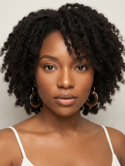 A close-up portrait of a woman with voluminous, tightly curled black hair, wearing large gold hoop earrings and a white spaghetti-strap top against a plain light gray background.