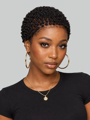 A woman with short, tightly curled black hair wearing large gold hoop earrings, a delicate gold necklace with a round pendant, and a black t-shirt, set against a solid gray background.