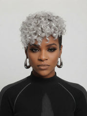Frontal portrait of a model in a silver-grey afro-textured pixie wig, highlighting the symmetrical curl pattern and fullness around the face. Her dramatic smoky eye makeup and nose piercing complement the edgy, confident vibe. The wig’s short length and tight coils create a striking contrast against her dark skin tone and minimalist black top.