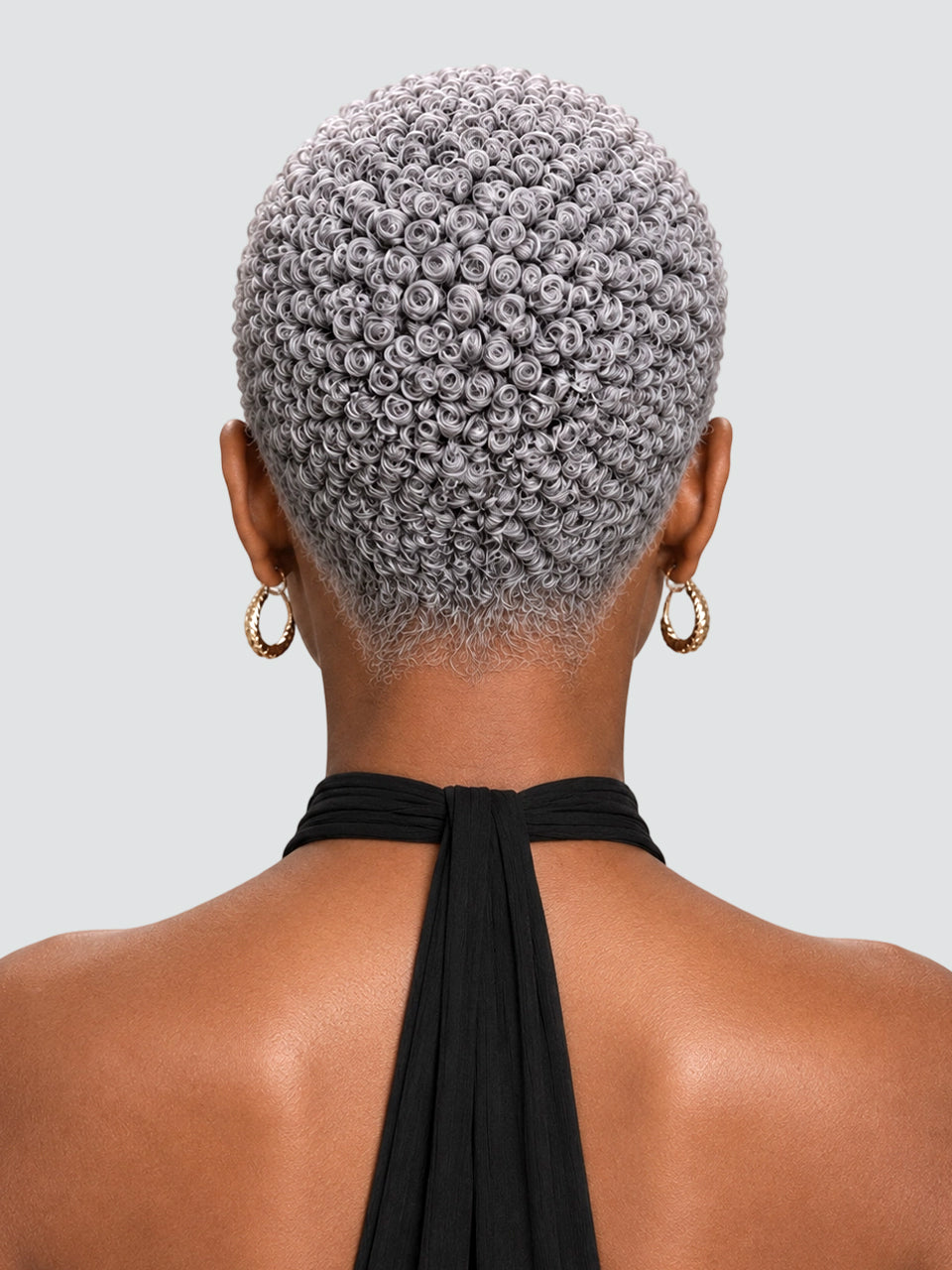 A woman in profile with a short, curly red hairstyle and shaved side details, wearing gold hoop earrings and a cream halter top against a light gray background.