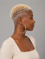 A woman in profile wearing large gold hoop earrings with pearl-like beaded detailing, a short textured blonde haircut, and a cream ribbed top against a solid gray background.