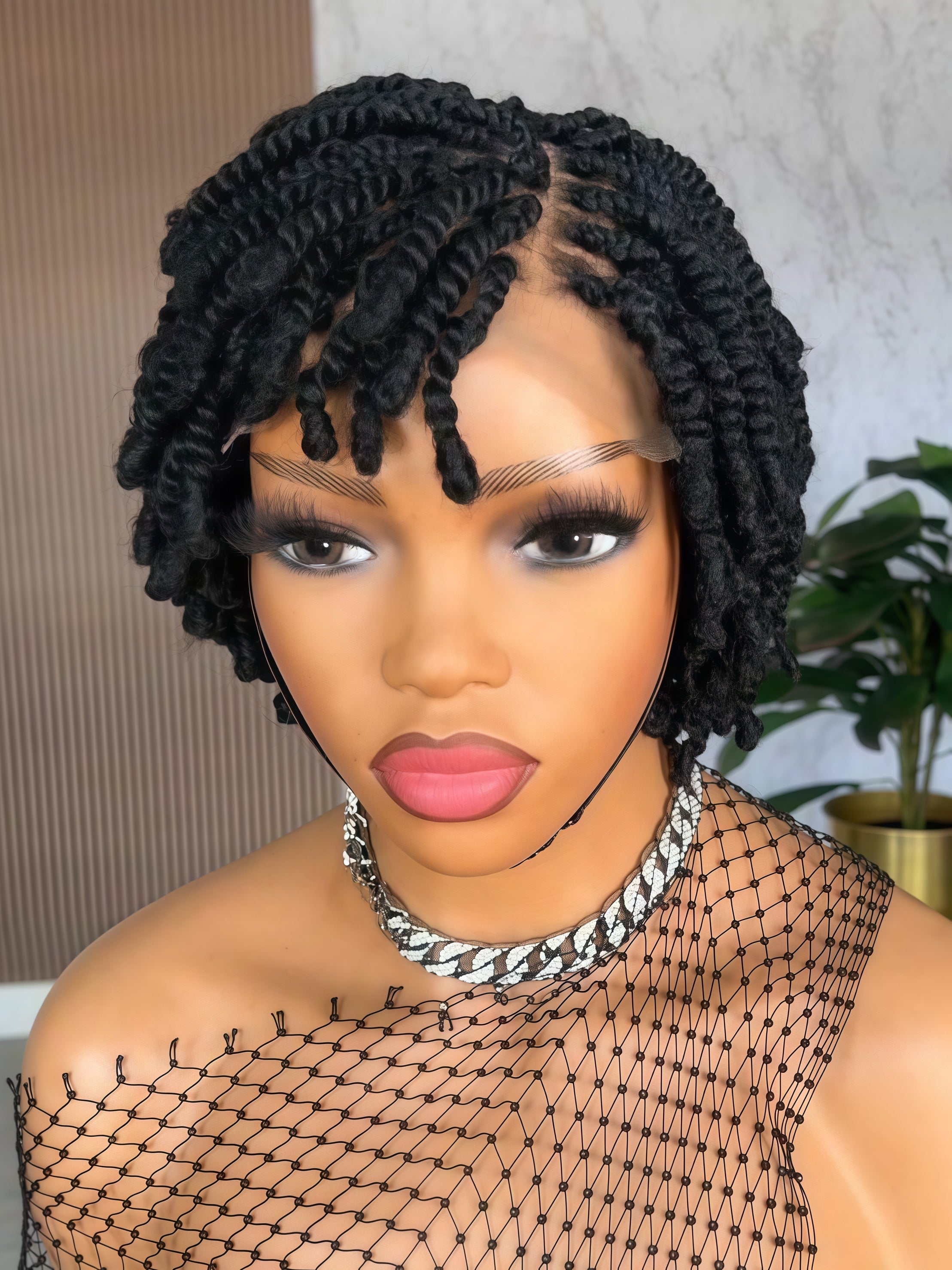 A close-up of a person wearing a short, curly black wig with a lace front and side parting, styled with dramatic eye makeup, pink lipstick, and a chunky silver chain necklace. The background shows an indoor setting with a textured wall and a potted plant.