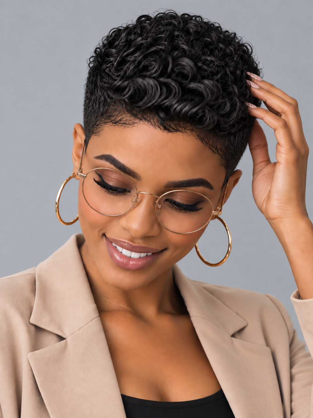 A woman with short, curly black hair wearing round gold-framed glasses, large gold hoop earrings, and a beige blazer, posing against a solid gray background.