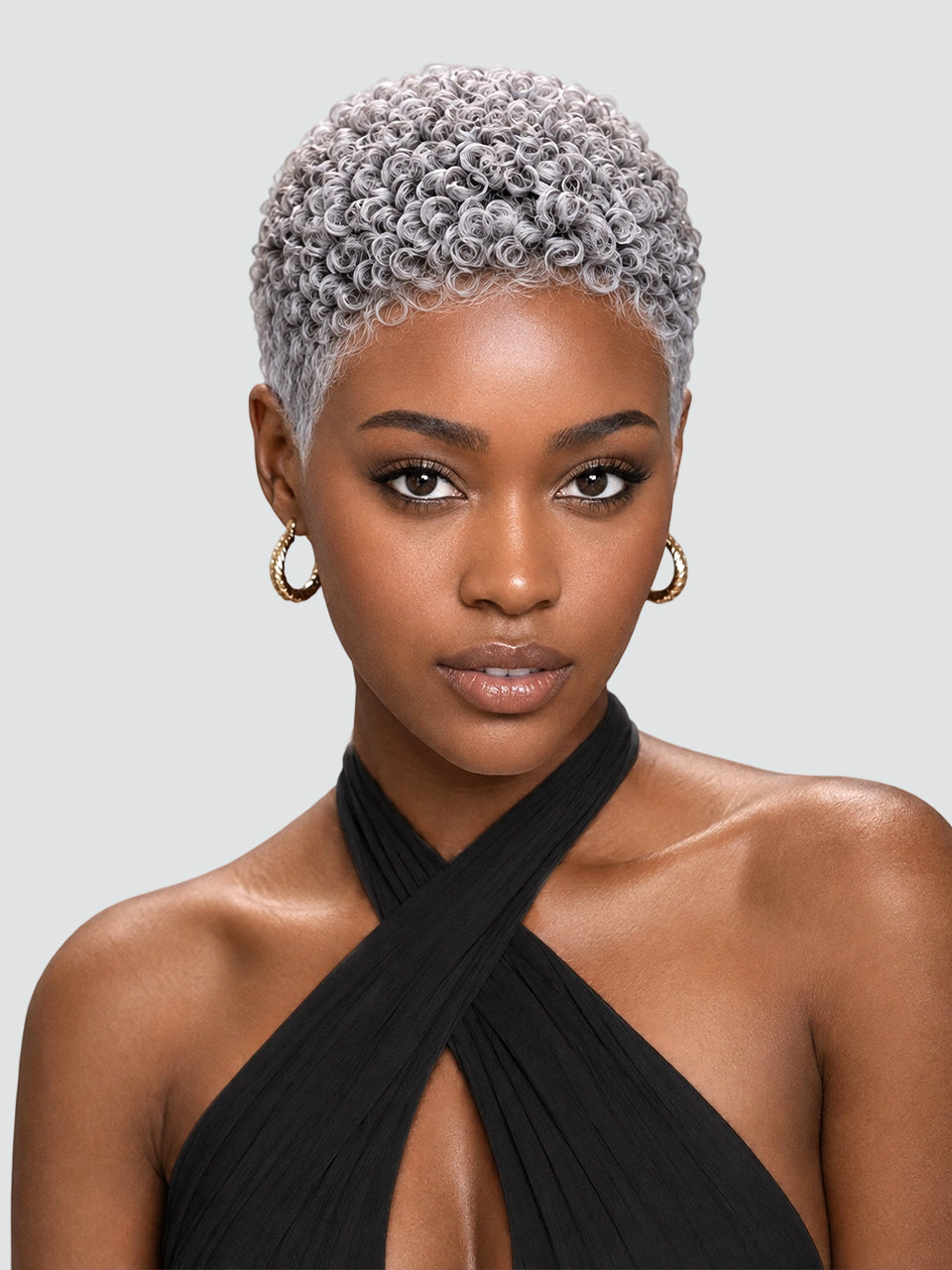 A close-up portrait of a model with a short, tightly curled platinum-blonde wig, wearing gold hoop earrings and a black halter top against a light gray background.
