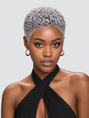 A close-up portrait of a model with a short, tightly curled platinum-blonde wig, wearing gold hoop earrings and a black halter top against a light gray background.