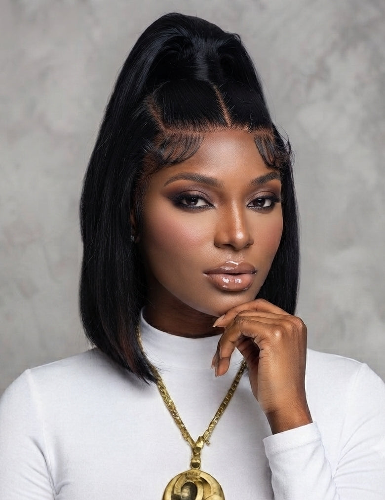 A woman with a sleek black high-top wig and bangs, wearing a white mock-neck top and a gold pendant necklace, posing against a textured gray background.