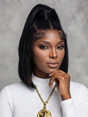 A woman with a sleek black high-top wig and bangs, wearing a white mock-neck top and a gold pendant necklace, posing against a textured gray background.