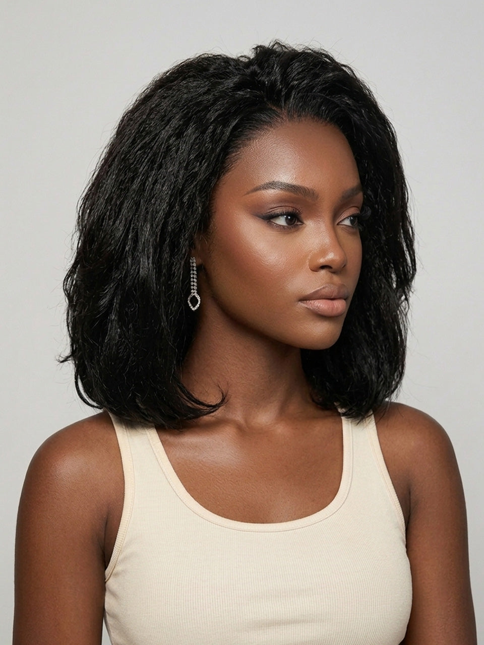 Three-quarter side profile of a woman in a black wavy bob wig, highlighting the gentle curl pattern and smooth transition from crown to ends. Her subtle makeup and dangling crystal earrings complement the sophisticated vibe. The image captures the realistic movement and shine of the hair against a neutral background.