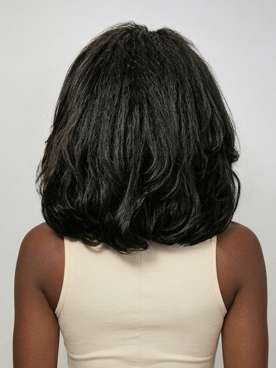 Back view of a model wearing a black medium-length curly wig, showcasing the voluminous, soft waves cascading down to the shoulders. The texture appears rich and bouncy, with natural-looking density that frames the neck elegantly. She pairs it with a simple beige tank top for a clean, minimalist aesthetic.