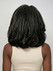 Back view of a model wearing a black medium-length curly wig, showcasing the voluminous, soft waves cascading down to the shoulders. The texture appears rich and bouncy, with natural-looking density that frames the neck elegantly. She pairs it with a simple beige tank top for a clean, minimalist aesthetic.