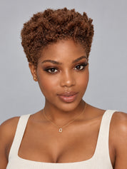 A woman with a short, curly brown wig or hairpiece shown from the shoulders up against a plain light gray studio background. She is wearing a white ribbed tank top and a delicate gold necklace.