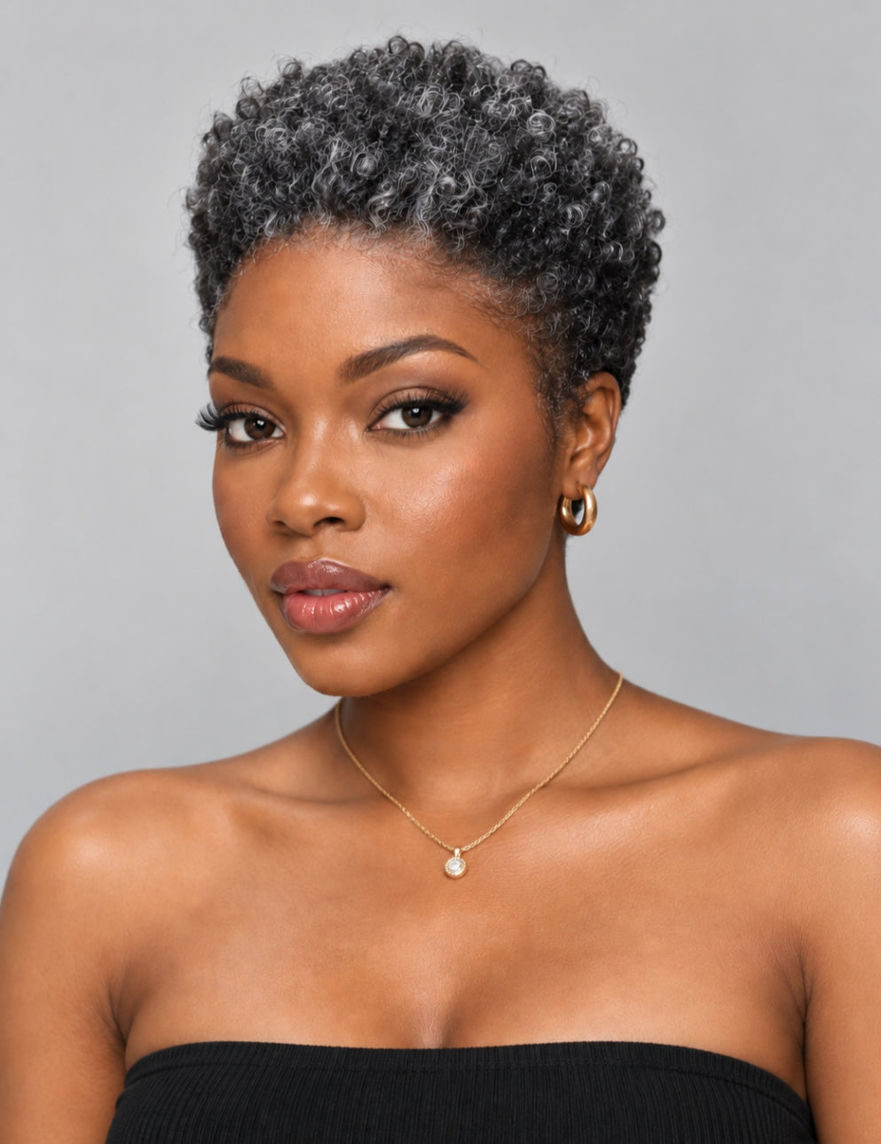 A woman with a short, curly gray wig or hairpiece, wearing a black strapless top, gold hoop earrings, and a delicate gold necklace with a pearl-like pendant, set against a solid gray background.