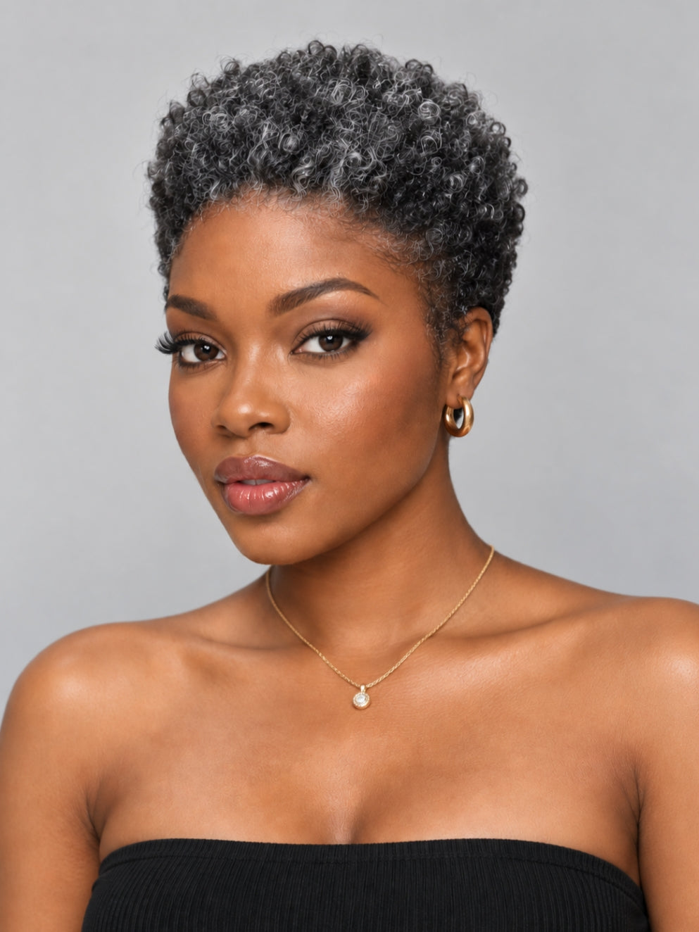 A woman with a short, curly gray wig or hairpiece, wearing a black strapless top, gold hoop earrings, and a delicate gold necklace with a pearl-like pendant, set against a solid gray background.