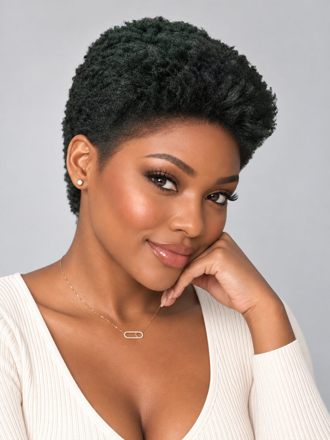 A woman with a short, voluminous dark green-black textured wig, wearing a cream ribbed top and a delicate gold necklace, posing against a solid gray background.