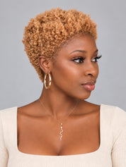 A woman with a short, tightly curled natural-looking hair extension or wig in a warm golden-blonde shade, styled with gold hoop earrings and a silver necklace, wearing a cream ribbed top against a solid gray background.