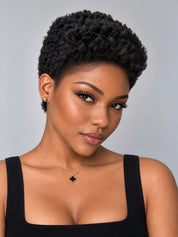 A close-up portrait of a woman with short, curly black hair, wearing a black ribbed tank top, a delicate gold necklace with a black clover pendant, and a small gold stud earring, set against a solid light gray background.