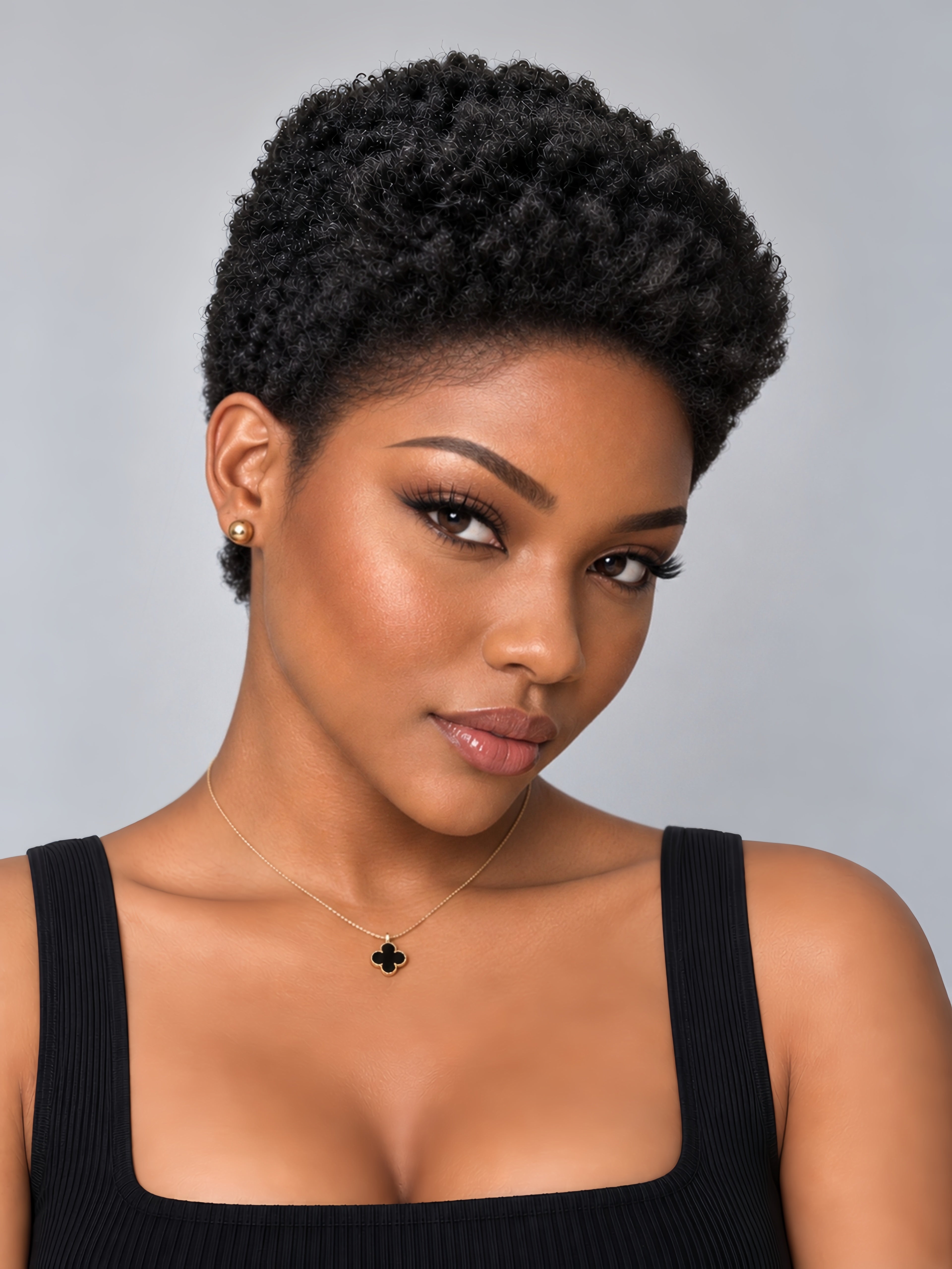 A close-up portrait of a woman with short, curly black hair, wearing a black ribbed tank top, a delicate gold necklace with a black clover pendant, and a small gold stud earring, set against a solid light gray background.