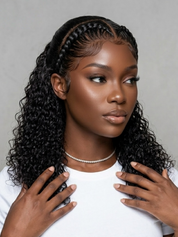 A woman with long, dark, curly hair styled with a center part and braided detailing along the crown, wearing a white t-shirt and a delicate pearl necklace against a neutral gray background.