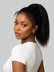 Back view of a woman with a short, curly, textured wig in dark brown with warm caramel highlights, paired with gold hoop earrings and a white spaghetti strap top, set against a solid gray background.