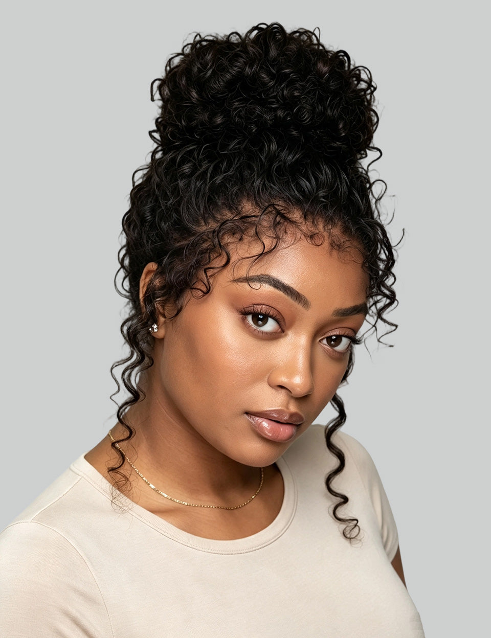 A woman with a high-volume, curly black wig or hairpiece styled in a voluminous updo with loose curls framing her face, wearing a light beige top against a solid gray background.