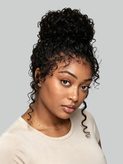 A woman with a high-volume, curly black wig or hairpiece styled in a voluminous updo with loose curls framing her face, wearing a light beige top against a solid gray background.