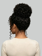 Back view of a person with a voluminous, high-volume curly updo hairstyle, wearing a cream-colored long-sleeve top against a solid gray background.