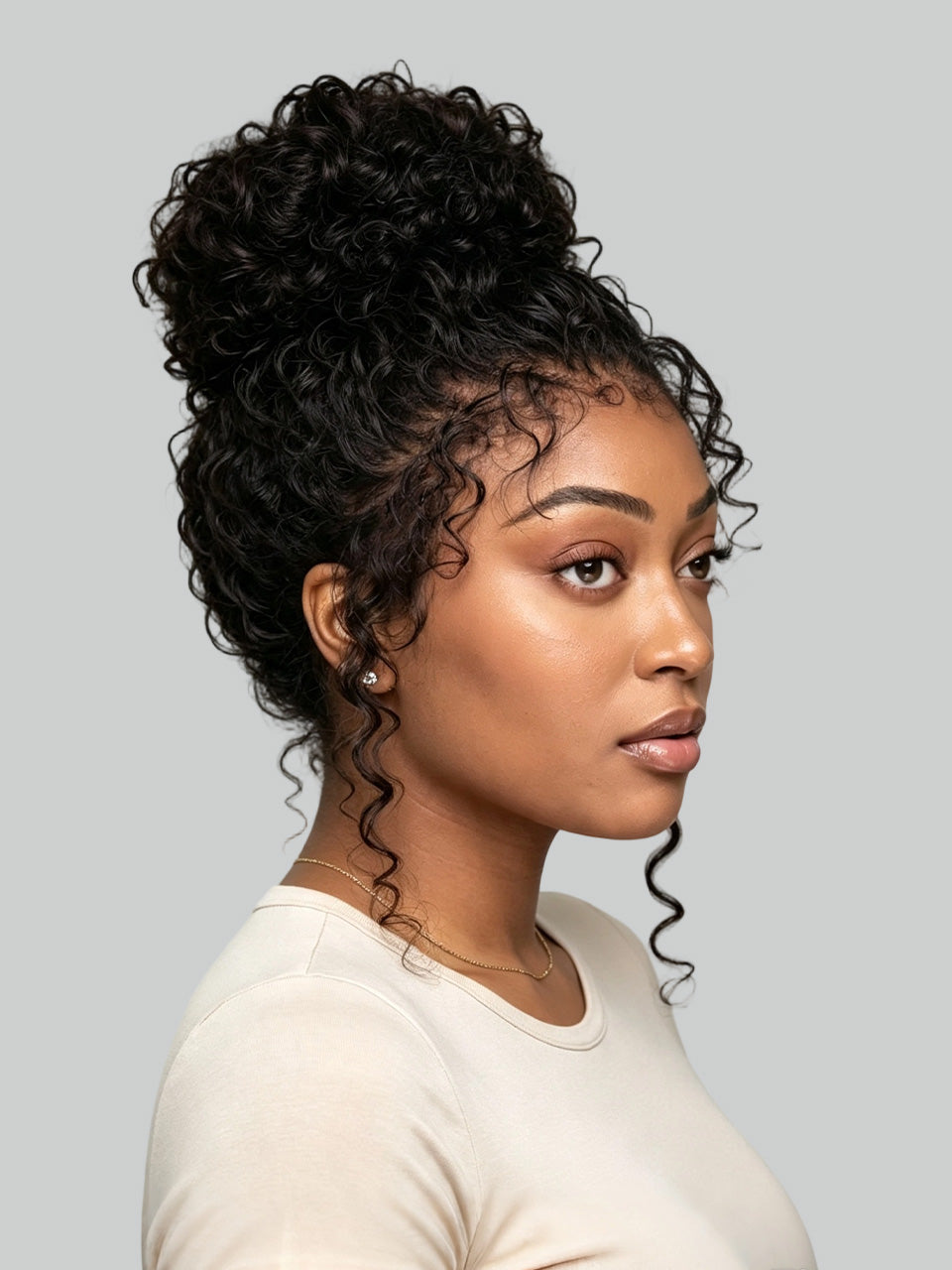 A woman shown in profile wearing a voluminous, curly black hairpiece or wig styled in a high bun with loose curls hanging near the shoulders, against a solid light gray background.