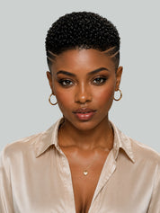 A close-up portrait of a woman with a short, tightly curled black wig, styled with a side part and gold hoop earrings, wearing a champagne satin shirt against a solid gray background.