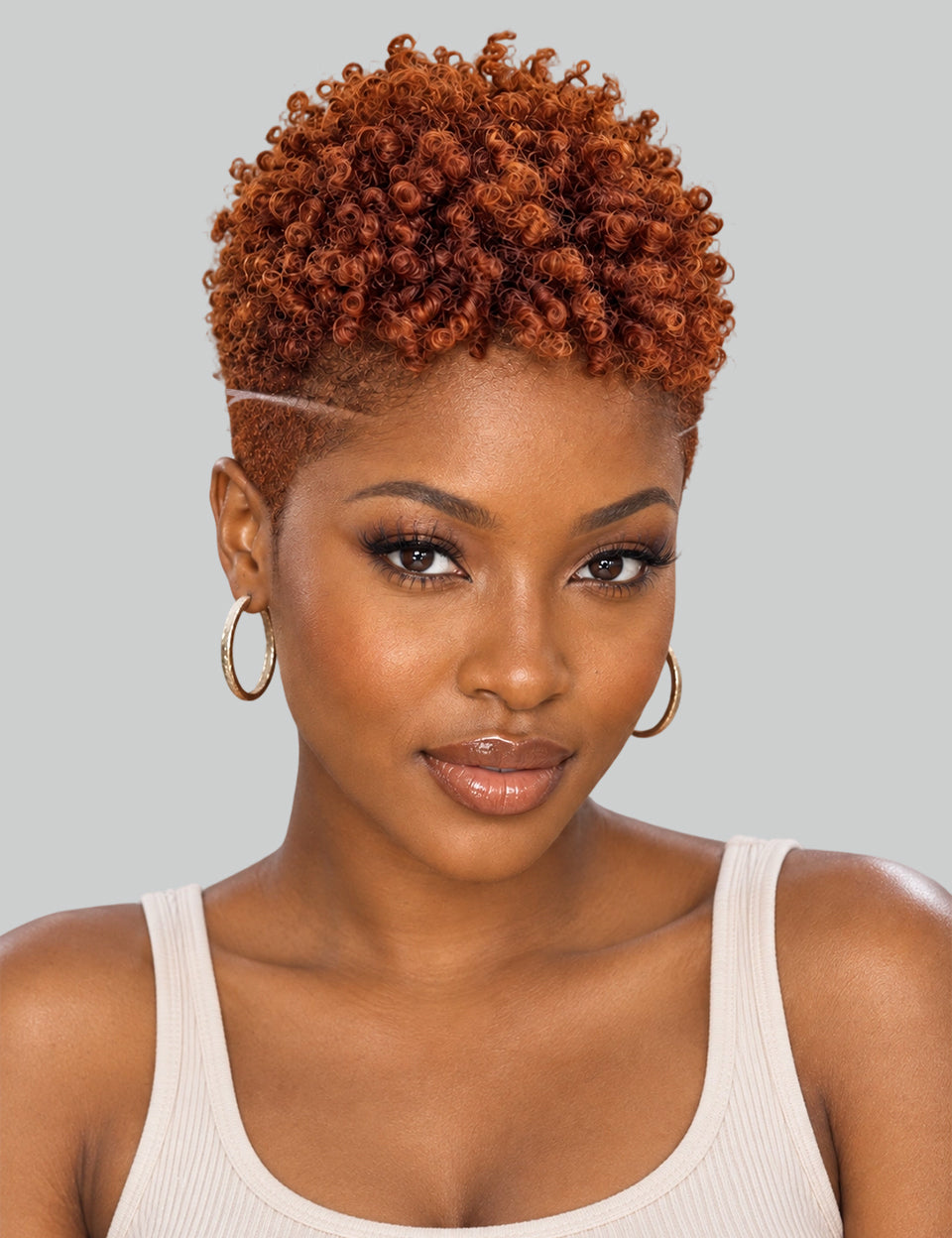 A close-up portrait of a woman with a short, tightly curled, textured wig in a warm coppery orange-brown shade. The wig is styled with a high side part and a thin white line accent across the forehead, paired with gold hoop earrings and a white ribbed tank top, set against a solid light gray background.