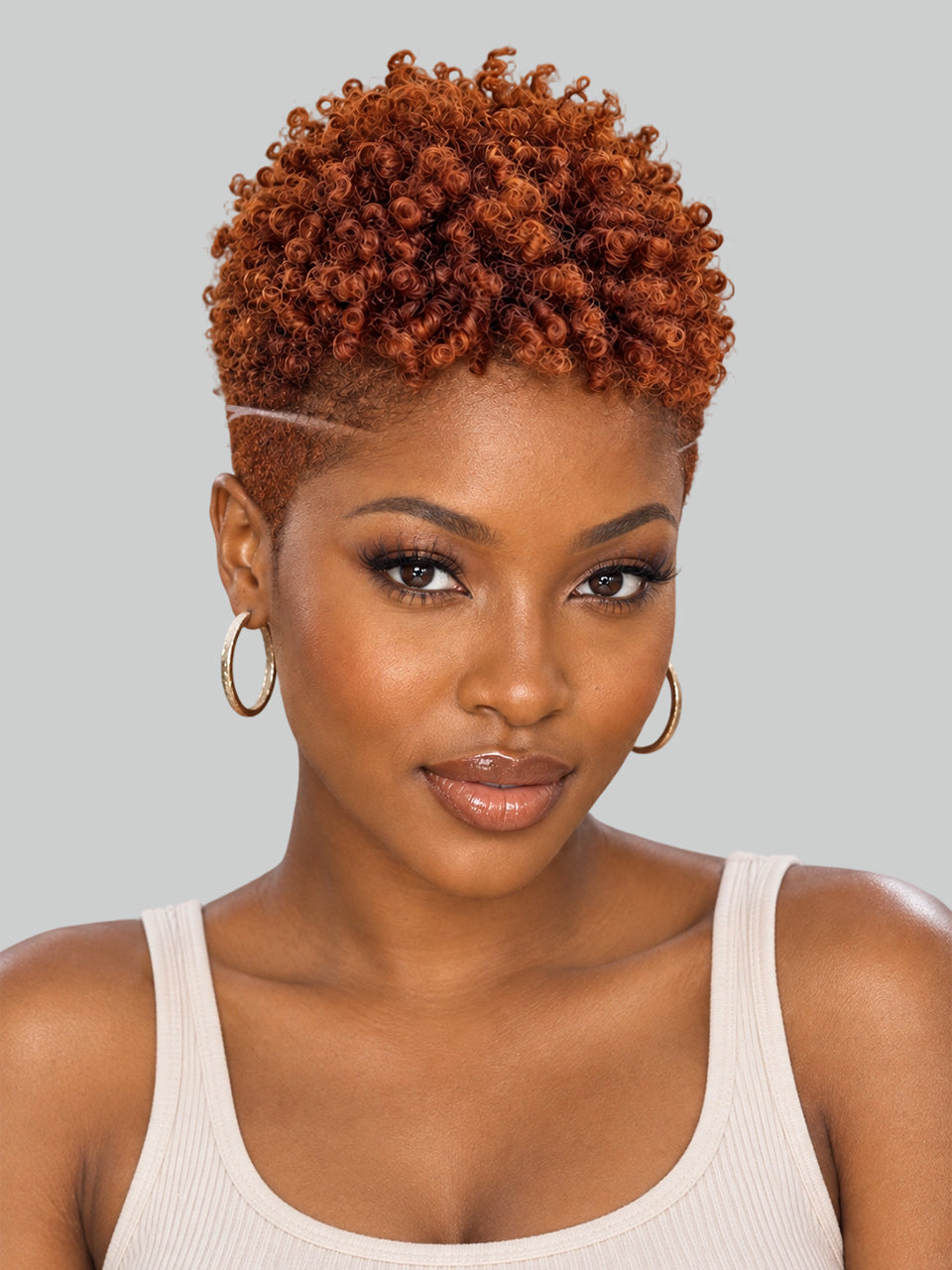A close-up portrait of a woman with a short, tightly curled, textured wig in a warm coppery orange-brown shade. The wig is styled with a high side part and a thin white line accent across the forehead, paired with gold hoop earrings and a white ribbed tank top, set against a solid light gray background.