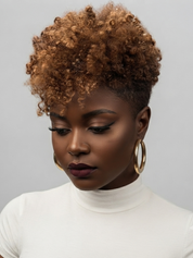 A close-up portrait of a woman with a short, voluminous, tightly curled afro hairstyle. She is wearing a white turtleneck top and large gold hoop earrings, looking down against a solid light gray background.